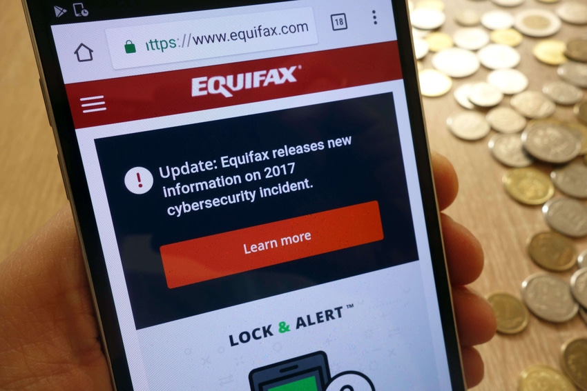 A notice about the Equifax breach from 2017 appears on the screen of a smartphone that is held up in front of a pile of coins