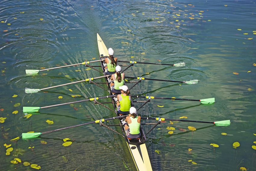 Photo of a team of four female rowers sculling down a leaf-littered river