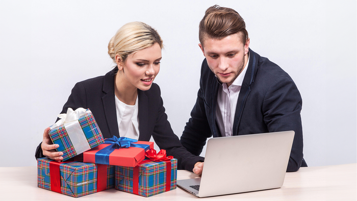 Businesswoman with ponytail in black suit and businessman in black suit with laptop sit at office desk with stack of presents, look at laptop