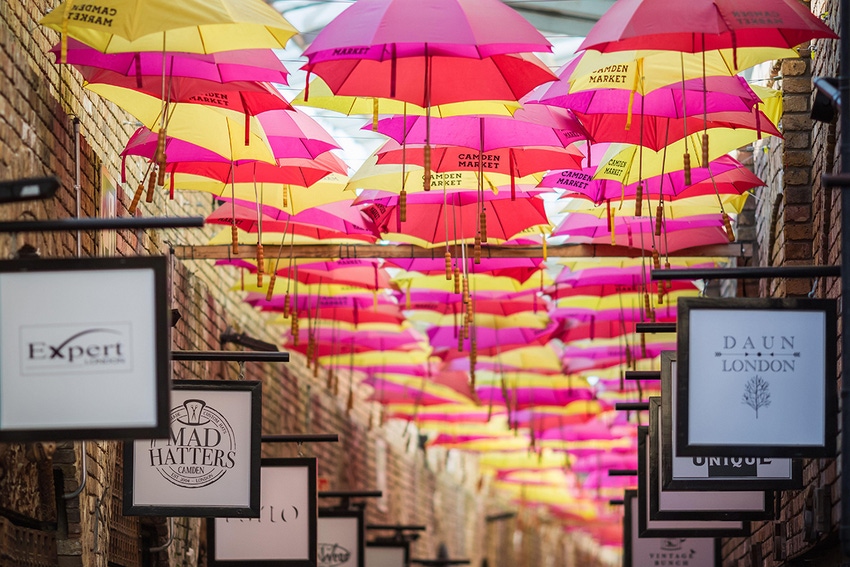 Pink and yellow umbrellas hang above business signs in the alley in Camden stables market