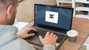 A man with his hands on the keyboard of a laptop, the display showing a pop-up featuring skull and bones reading "you've been hacked"