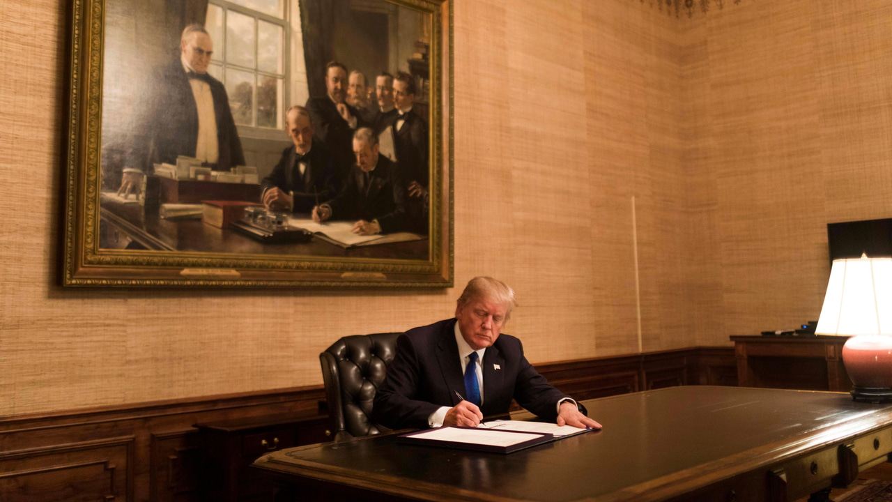 President Trump signing a document at a large desk in front of a painting President Trump signing a document at a large desk in front of a painting
