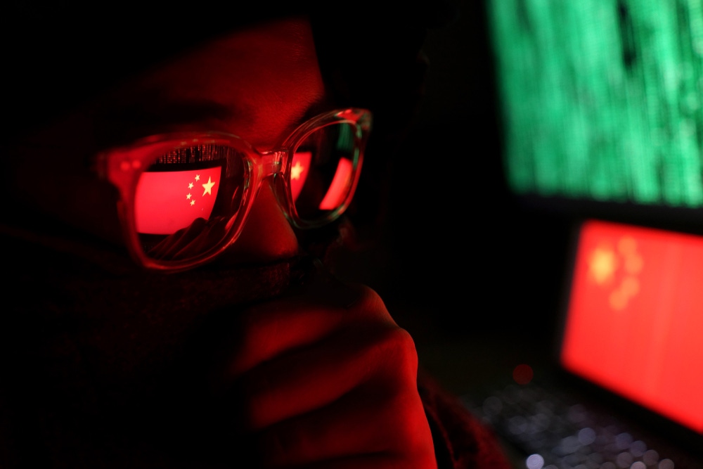 Hacker in a dark wearing hoody and cap sitting in front of computer with digital China flag background and binary code Hacker in a dark wearing hoody and cap sitting in front of computer with digital China flag background and binary code