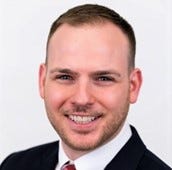 David Homovich is wearing a white collared shirt and a dark jacket, and has a short beard and short haircut David Homovich is wearing a white collared shirt and a dark jacket, and has a short beard and short haircut