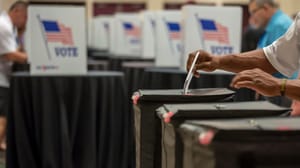 A person putting their ballot in a ballot box in front of a bunch of other voting booths A person putting their ballot in a ballot box in front of a bunch of other voting booths