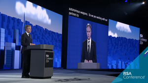 US Secretary of State Antony Blinken stands at a lectern on a large stage with projection screens everywhere.