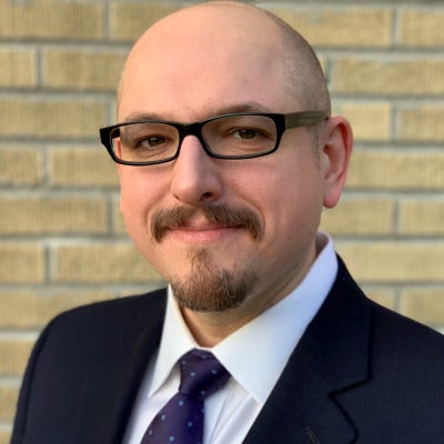 Daniel Dobrygowski has glasses, a shaved head, and a brown Van Dyke beard and mustache. He's wearing a dark suit and tie.