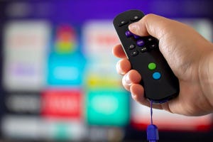 A person holding a Roku remote control up to a blurred TV in the background