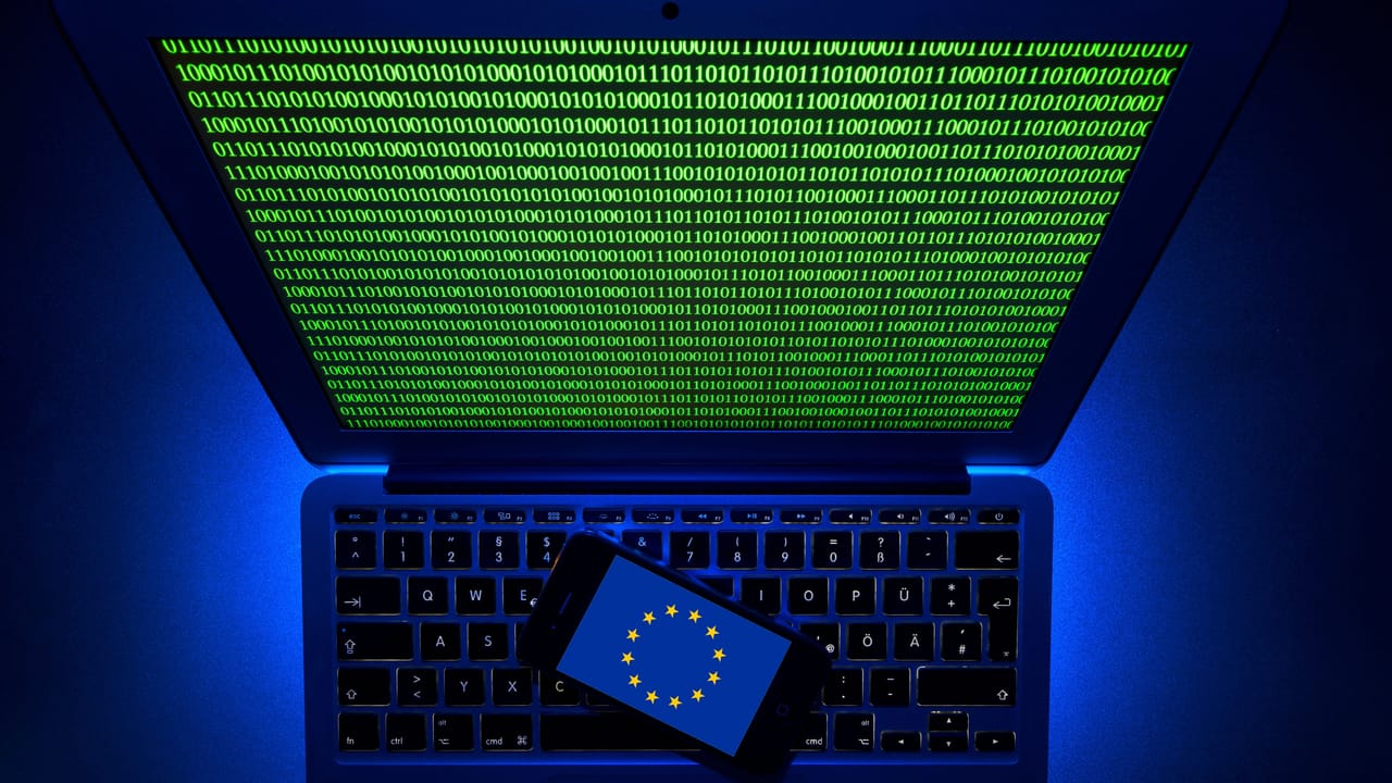 ヨーロッパの国旗が表示されたスマートフォンが、緑色のバイナリコードで埋め尽くされたモニターを備えたコンピュータキーボードの上に置かれている ヨーロッパの国旗が表示されたスマートフォンが、緑色のバイナリコードで埋め尽くされたモニターを備えたコンピュータキーボードの上に置かれている