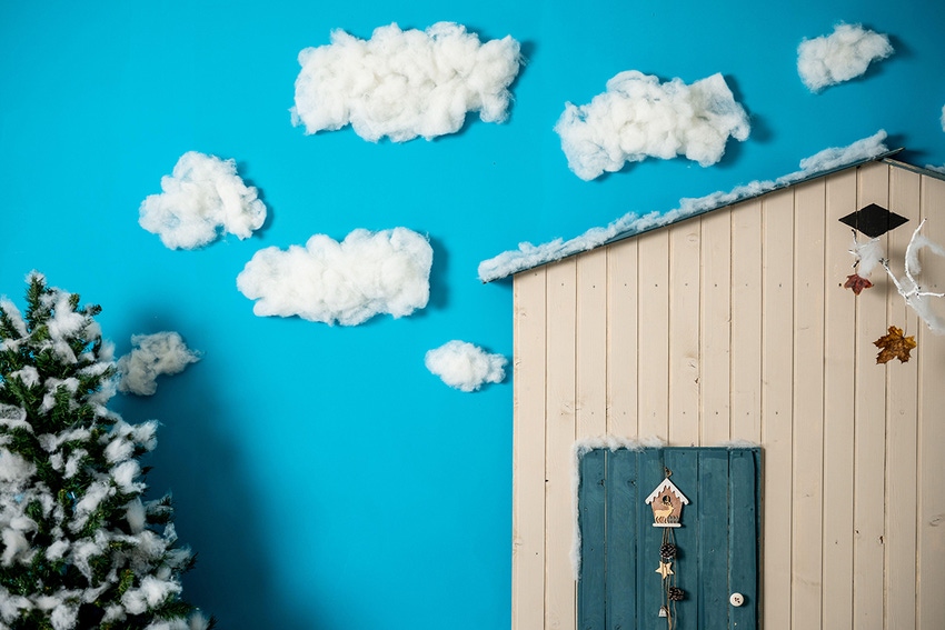 Diorama of a fake house with blue door, with fake cottony clouds and snow covering a fake pine tree