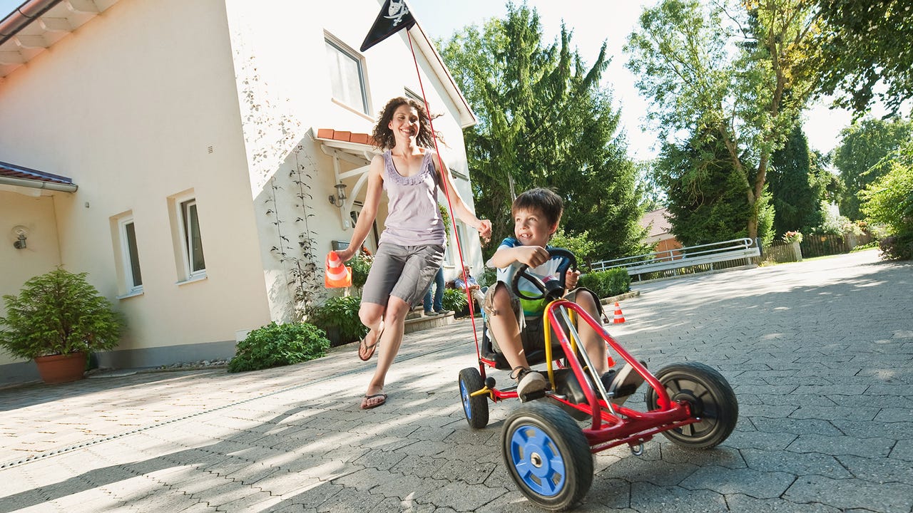 In Bavaria, boy driving pedal go-kart on a tiled driveway is chased by a running woman. Both are smiling. A house and trees are in the background.