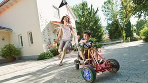 In Bavaria, boy driving pedal go-kart on a tiled driveway is chased by a running woman. Both are smiling. A house and trees are in the background. In Bavaria, boy driving pedal go-kart on a tiled driveway is chased by a running woman. Both are smiling. A house and trees are in the background.