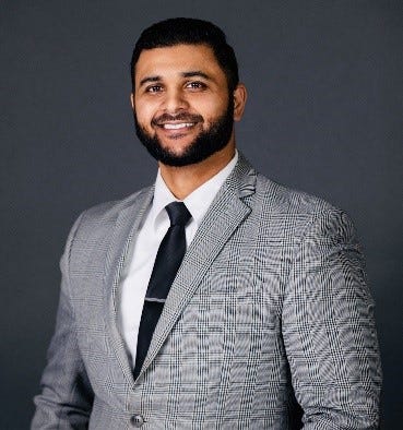 Nader Zaveri has a dark neatly-trimmed beard and is wearing a grey suit, white shirt, and a dark tie.