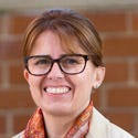 Kris Lovejoy wears tortoiseshell-framed glasses and a scarf. She is standing in front of a brick wall.