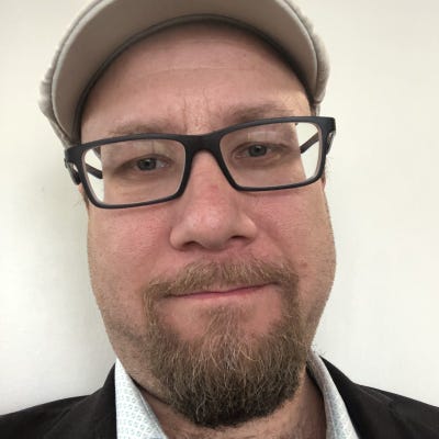 Luke Smith, Senior Director of Solution Engineering at Asimily, wears dark-framed glasses, a Van Dyke beard, and a flat cap