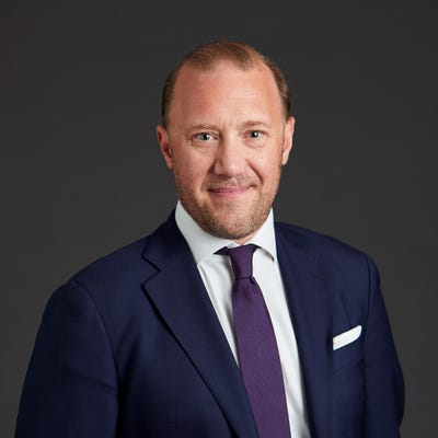 David Dunn has blue eyes and buzzed red hair and beard stubble; he wears a dark jacket and tie with white shirt