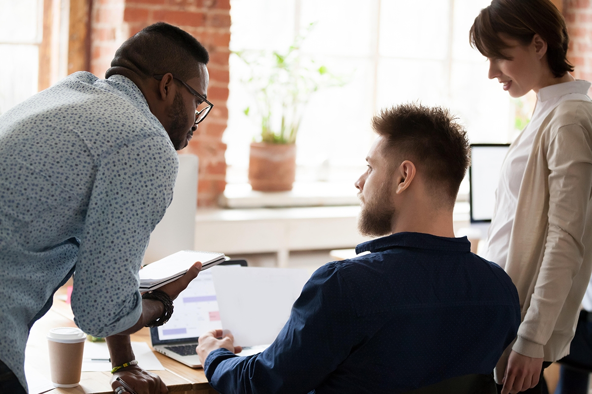 Three young colleagues in business casual clothes gather around a computer to discuss a solution