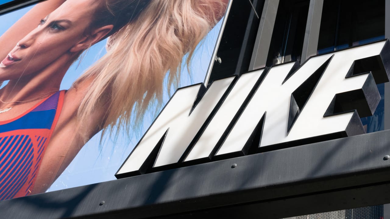 オレンジと青のシャツを着た金髪ポニーテールの女性が写る看板の横に、白いブロック体で「Nike」という文字 オレンジと青のシャツを着た金髪ポニーテールの女性が写る看板の横に、白いブロック体で「Nike」という文字