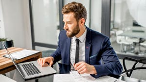 A bearded man with auburn hair and wearing a suit works on a laptop and paperwork at his desk