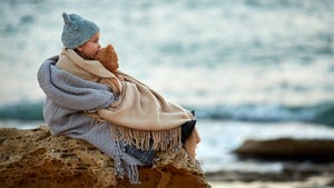 A little girl with a teddy bear stares out to sea, sitting on a stone on a cold beach, wrapped in blankets