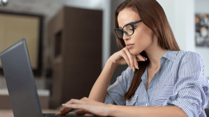 Dark-haired woman with long hair and black glasses in pin-striped shirt sitting at a computer with one hand on the keyboard abd one under her chin Dark-haired woman with long hair and black glasses in pin-striped shirt sitting at a computer with one hand on the keyboard abd one under her chin