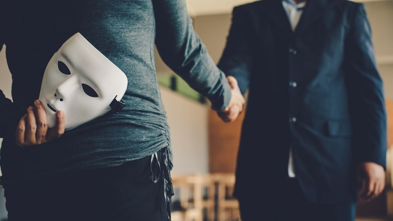 Two people in dark blue suits shaking hands, but one person is hiding a white mask behind their back. Two people in dark blue suits shaking hands, but one person is hiding a white mask behind their back.
