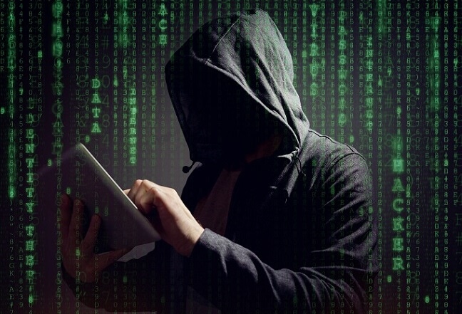 Image shows a dark figure in a hooded sweatshirt typing at a computer in front of lines of code on a computer screen
