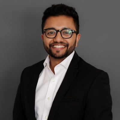 Nebu Varghese has black hair and wears dark-framed glasses, a short beard, and a black jacket over an open collar oxford Nebu Varghese has black hair and wears dark-framed glasses, a short beard, and a black jacket over an open collar oxford