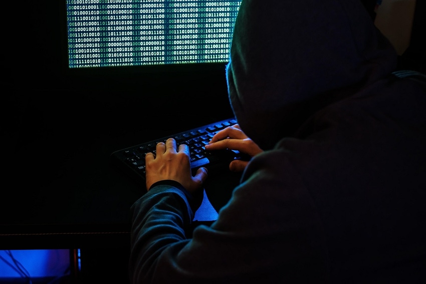 Image shows a dark figure in a hooded sweatshirt typing at a computer in front of lines of code on a computer screen