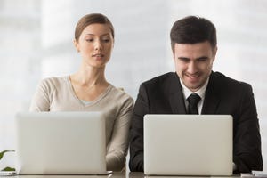 Female curious corporate spy sneaking a look at male colleague's laptop screen over his shoulder.