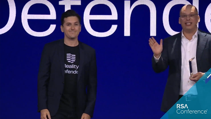 Reality Defender co-founder and CEO Ben Colman wears a company t-shirt and blazer onstage with host Hugh Thompson, who holds an award