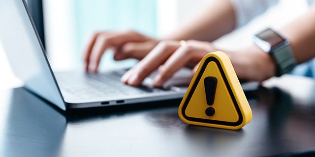 Photo of woman typing on laptop in the background while a yellow caution sign in the foreground warns of fraud