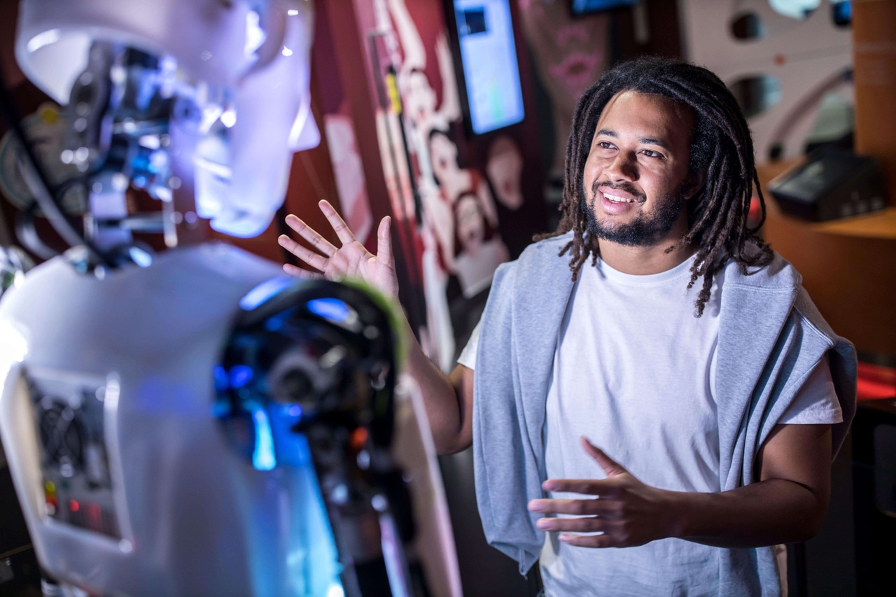 A man with locs and a short beard and mustache gestures to a white robot seen from behind A man with locs and a short beard and mustache gestures to a white robot seen from behind