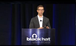 Scott Tenaglia, a brown-haired man wearing glasses and a suit, no tie, stands at a podium with Black Hat 2022 branding
