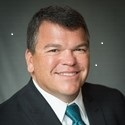 Brennan P. Baybeck, VP & CISO for Customer Services for Oracle, in a black suit with green tie. He has brown hair and eyes.
