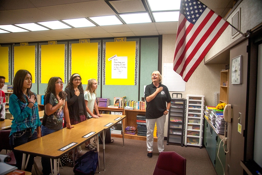A teacher and children in a classroom stand and put their right hand over their hearts to pledge allegiance to the US flag hanging in the corner