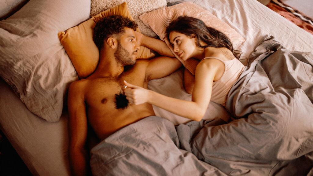 Nainen ja mies makaavat sängyssä ja katsovat toisiaan samalla kun nainen kutittaa miestä höyhenkutittimella