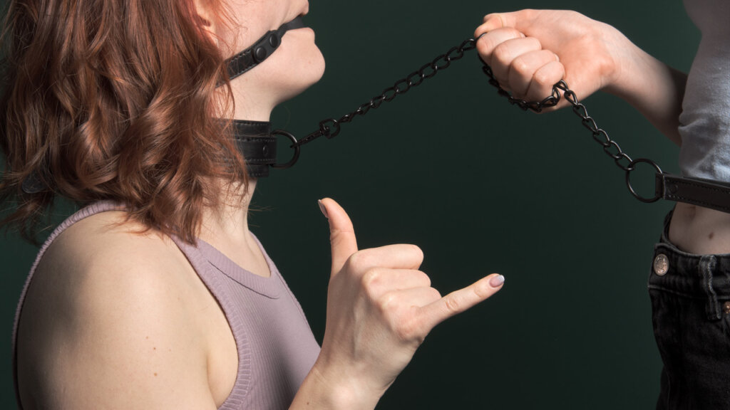 Kvinne med gagball og halsbånd viser et tegn med hendene 