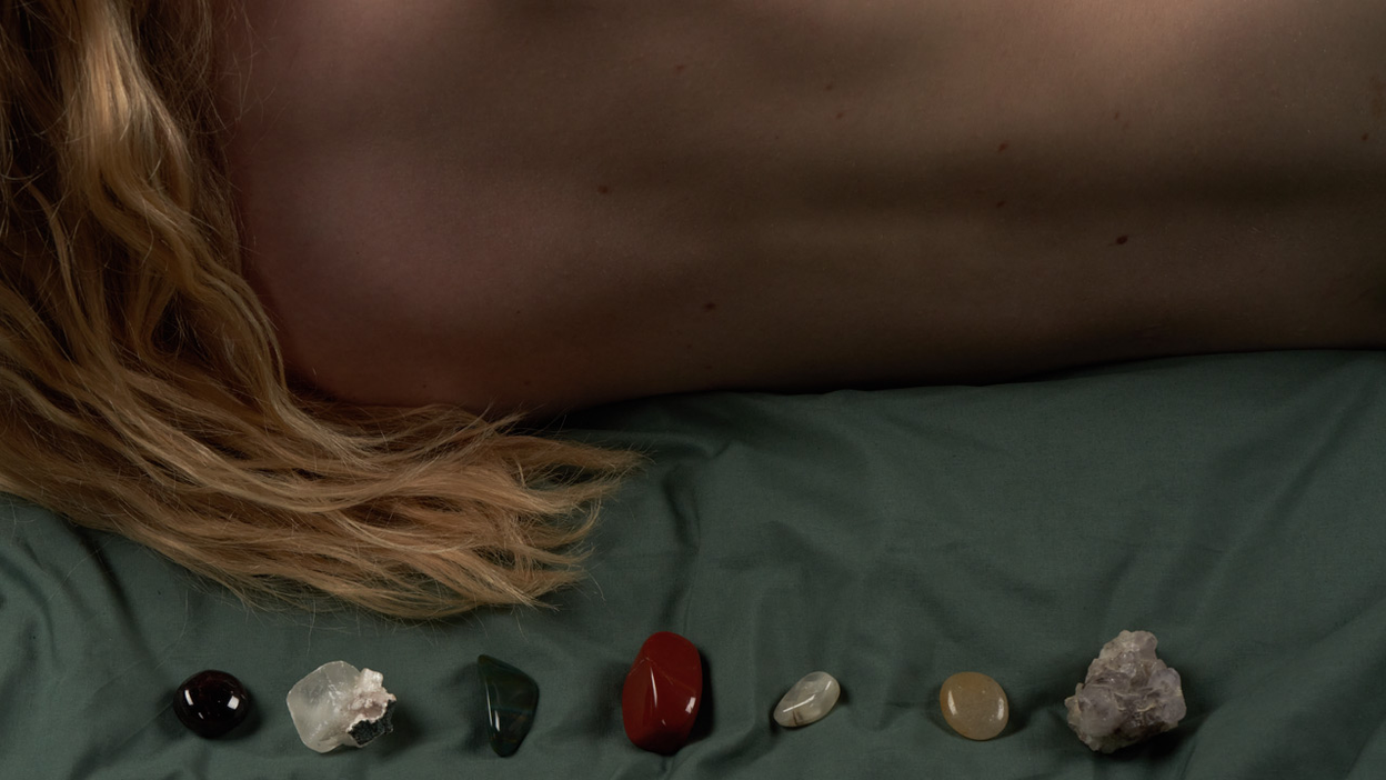 seven chakra crystals lined up on dark green sheet with a models bare back behind them