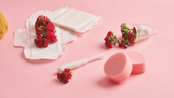 En tampon, bind, svampe, blomster og en menstruationskop ligger på en lyserød baggrund.
