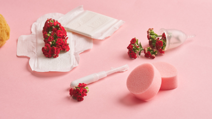 En tampon, bind, svampe, blomster og en menstruationskop ligger på en lyserød baggrund.