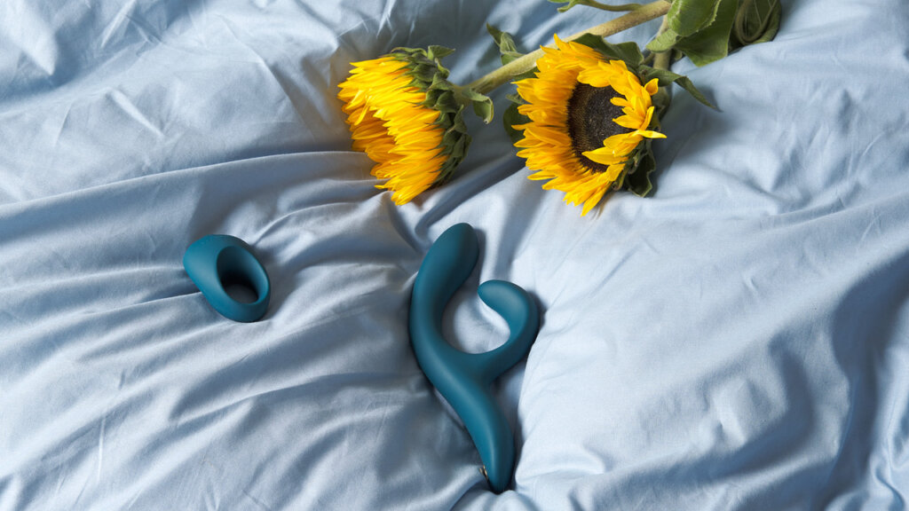 A rabbit vibrator and a cock ring laying on a light duvet cover beside two sunflowers