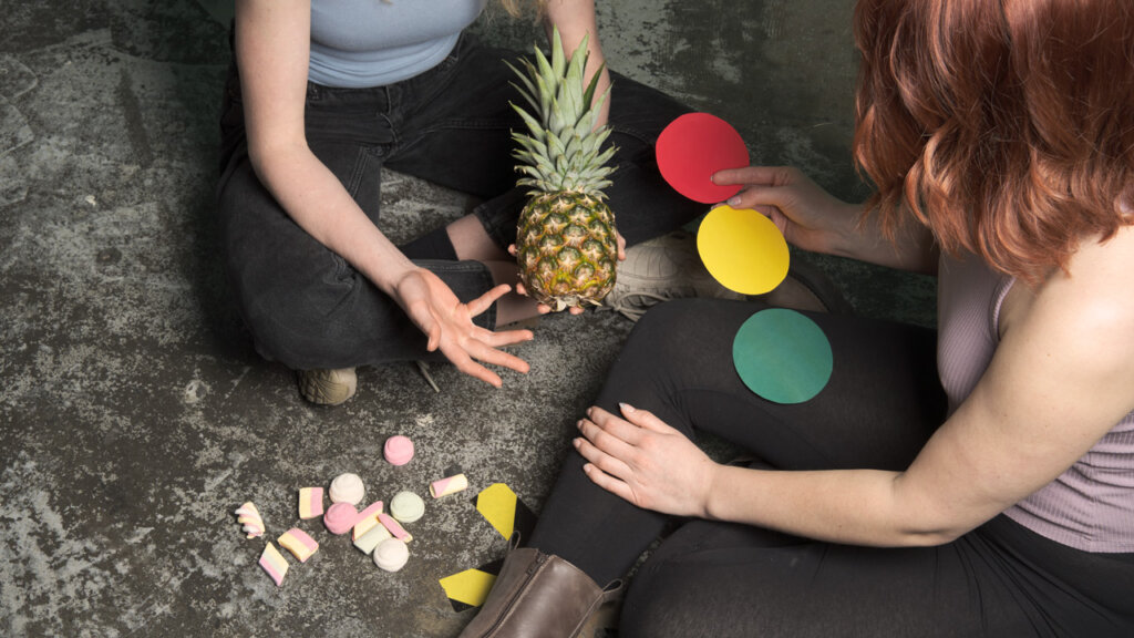 Twee vrouwen zitten met een ananas, marshmallows en drie stukjes gekleurd papier 