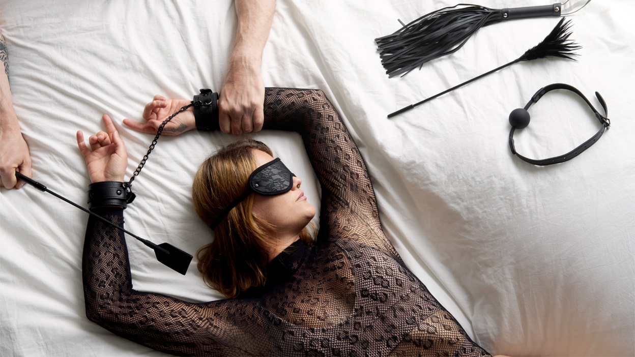 A person in lingerie lying on bed with hands cuffed above their head and a flogger, gag and feather beside them