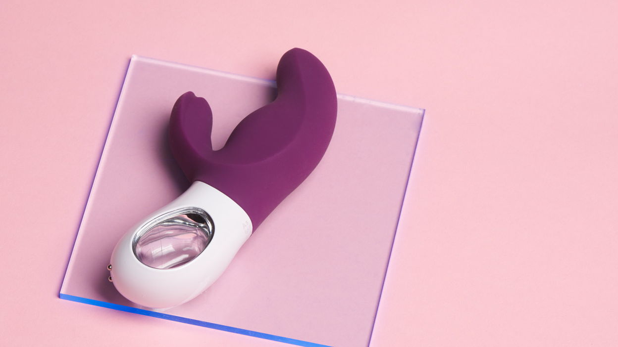 Ein lilaner und weißer Rabbit-Vibrator liegen auf einer Glasplatte auf einem rosa Hintergrund