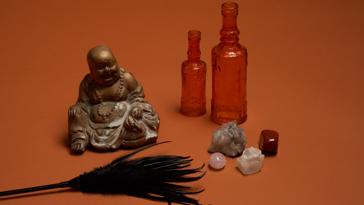 En kilefjær, en Buddha figur, to flasker og små krystaller står på en oransje bakgrunn