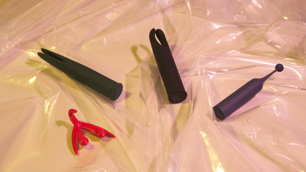 Tre klitoris vibratorer og en model af klitoris ligger på gennemsigtigt plastik på en beige baggrund