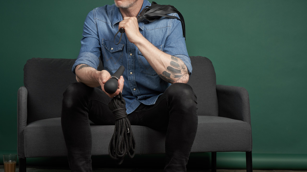 A person is sitting on a sofa with a bondage rope in their hand and a flogger on their shoulder