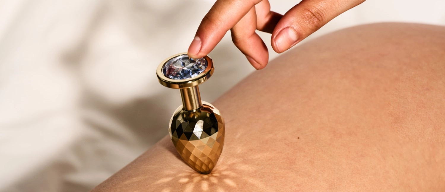 Een hand die een gouden buttplug met steentje tegen blote huid houdt.