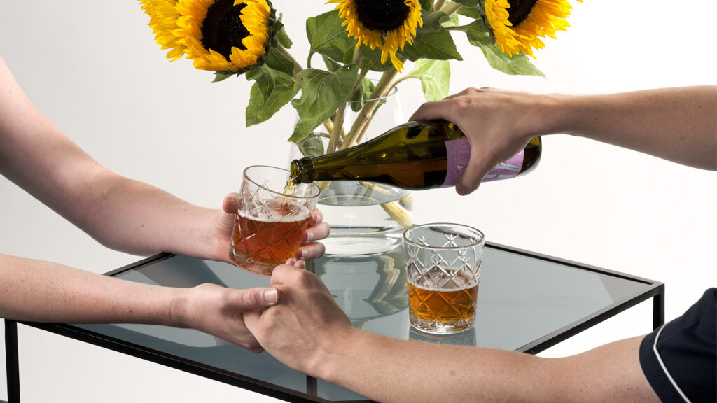 Två personer händerna över ett bord med två glas öl och en vas med solrosor